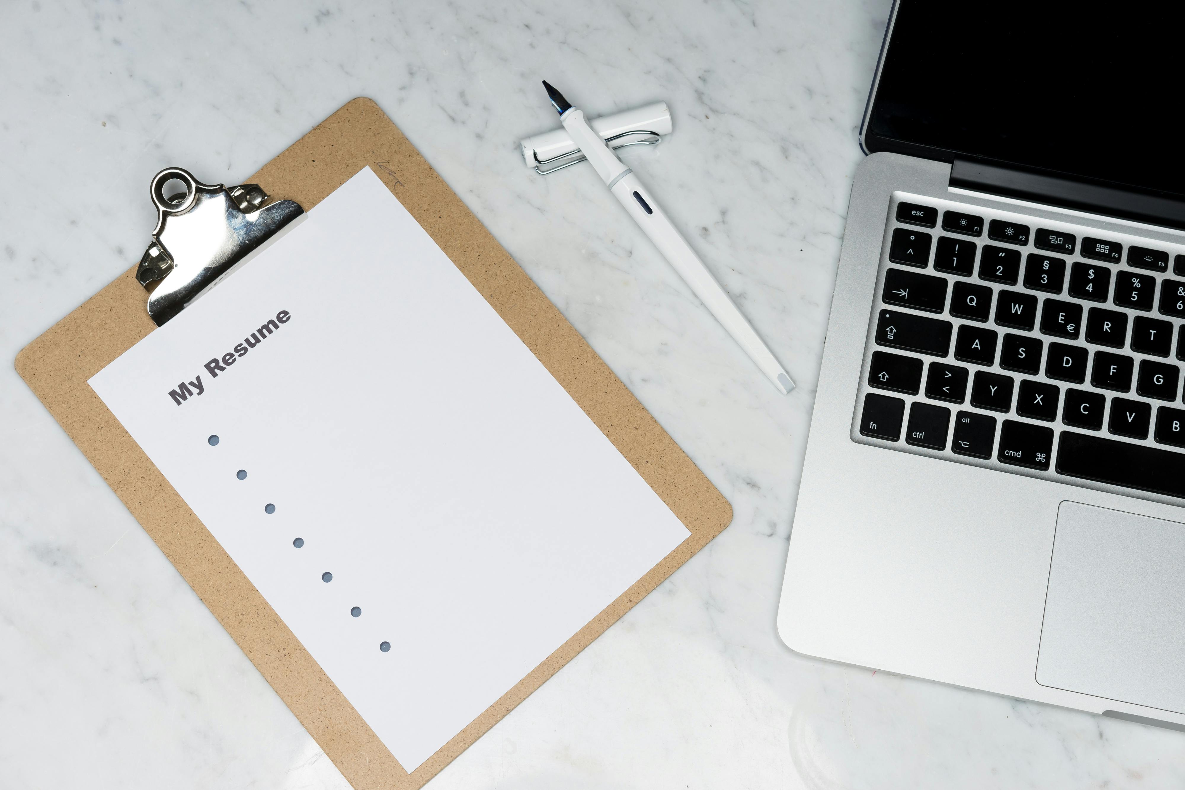 Resume on clipboard with pen and laptop on desk